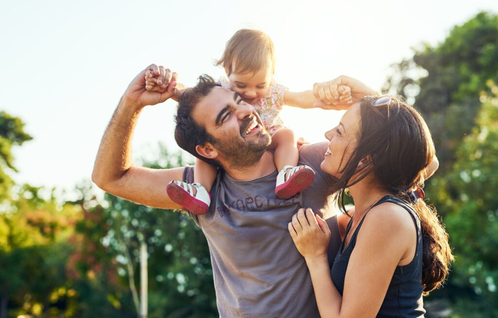 A young girl on top of her dads shoulders while he is standing next to a woman. They are all laughing.