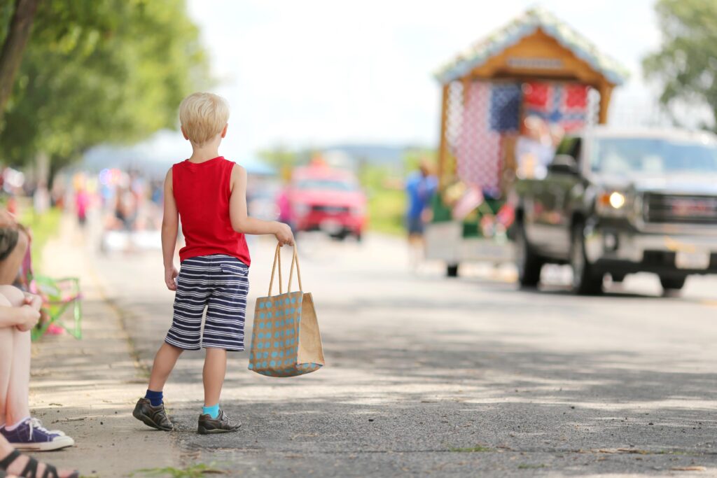 a child standing with a bag on the side of a parade.